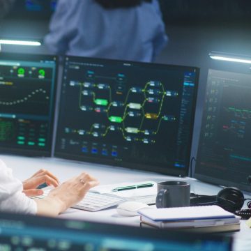 Female IT technical specialist works on computer with data server and blockchain network database in modern monitoring control room. Team of software engineers and big digital screens on background.